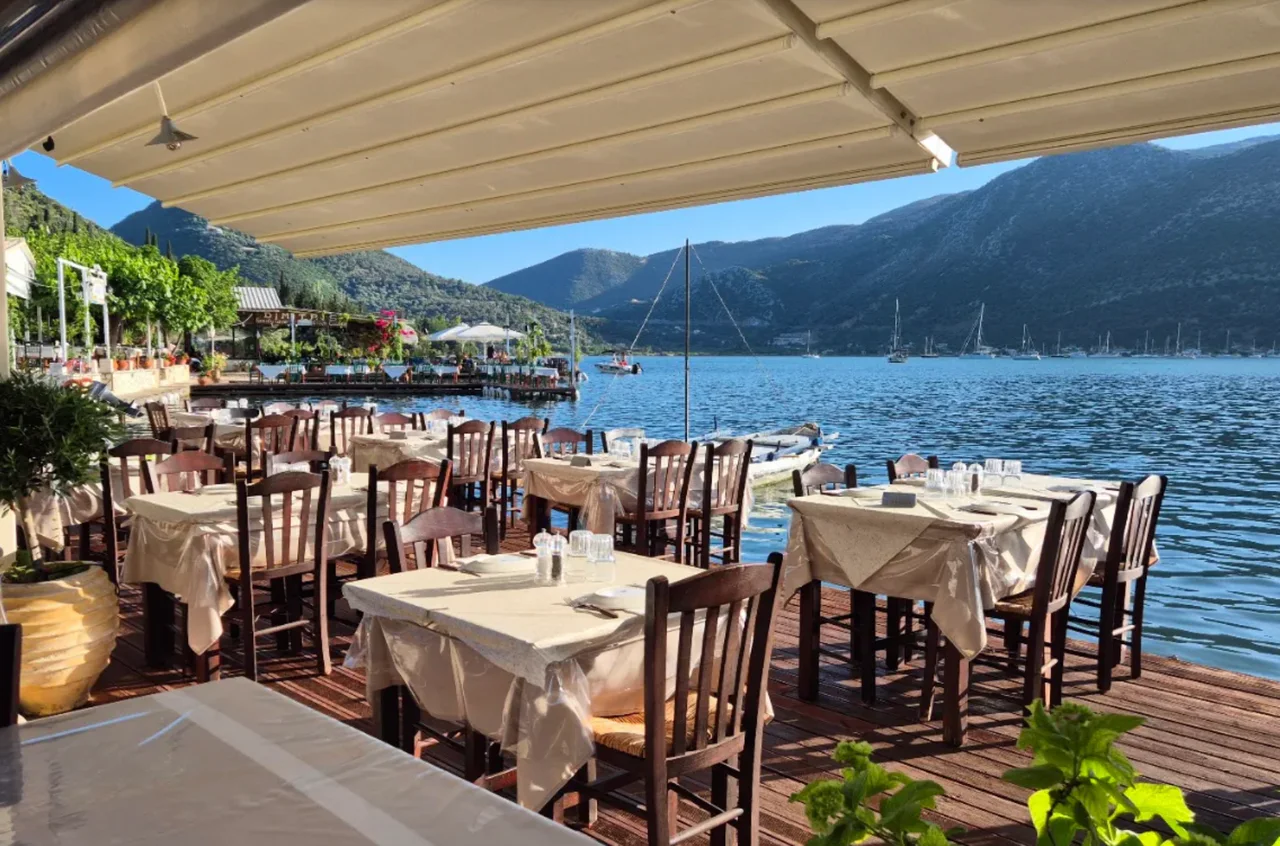 Waterside taverna tables beside Geni Bay, with sailing boats and green hills beyond